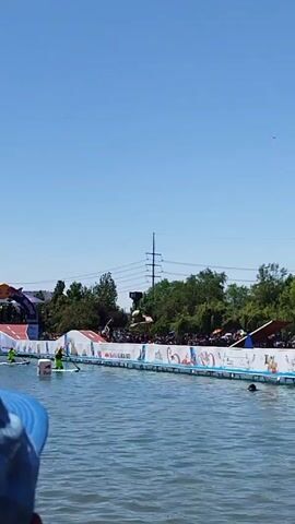 The video captures a water event on a clear day. A structure with advertising appears to be the main platform for competitors. Several people, likely participants or support staff, are in the water. In the background, a crowd is observing the event, with trees and power lines also visible. The video is a steady, distant shot of the scene.