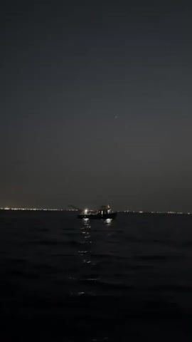 The video shows a wide shot of a dark body of water at night. A small boat with lights on is in the center of the frame, with a long pole or boom extending from its left side. In the distance, a faint line of lights suggests a shoreline or a city. The sky is dark and clear, with a few faint stars visible. The water is choppy, with reflections of the boat's lights on the surface. The camera is slightly tilted upwards, giving a sense of looking out over the water towards the horizon.