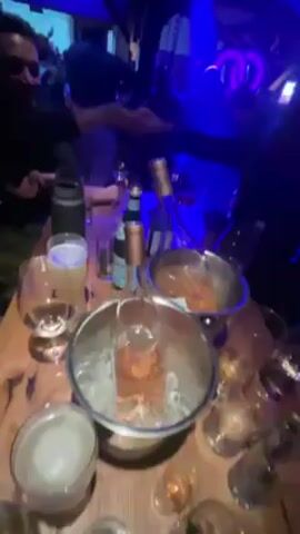 The video shows a group of people at a club or bar, likely celebrating.  The camera focuses on a table with multiple wine bottles in ice buckets, various glasses, and a bottle of sparkling water.  The table appears to be made of wood.  Behind the table, people are dancing and socializing in a dimly lit space.  A DJ or performer is visible on a stage under bright blue lights. A sign behind them reads "...LOOSER. W".  The architecture is characterized by exposed wooden beams, giving the space a rustic feel, possibly a converted barn or similar structure.  The lighting alternates between blue, purple and red.