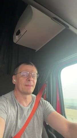 A man in a gray t-shirt and glasses sits in the driver's seat of a semi-truck, wearing a red seatbelt. He has wireless earbuds in his ears. The interior of the truck is visible, with a storage compartment above him. He sings with his mouth open wide. The exterior of the truck is obscured by a foggy or overcast sky.