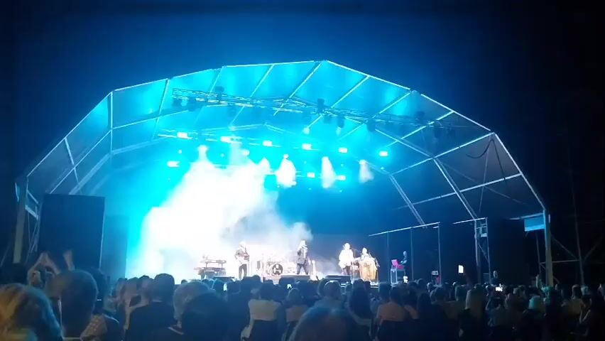 A band performs on a stage at night. The stage is covered by a large, white, geodesic-like tent structure. Blue spotlights illuminate the band and create smoke effects. A crowd of people watches the performance from the foreground. The shot is taken from behind the crowd, looking towards the stage.