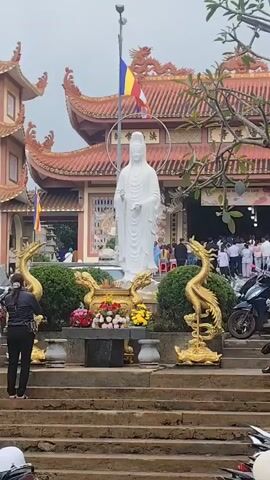 A bustling scene unfolds in front of a temple entrance. A large white statue of a deity stands prominently on the left, adorned with offerings of flowers at its base. Flanking the statue are decorative golden dragon and phoenix figures. A flight of stone stairs leads up to the temple's entrance, where numerous people, many dressed in white, are gathered. Some are standing, while others appear to be kneeling or bowing in prayer. Motorcycles are parked to the right of the entrance. Flags, including a Buddhist flag, are visible, adding to the ceremonial atmosphere. The temple itself features traditional Asian architecture with a red tiled roof.