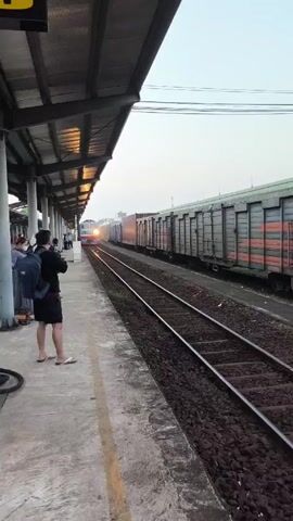 The video shows a train approaching a station platform. The train has a light blue and white engine and multiple cargo cars. Several people are standing on the platform, waiting for the train or watching it arrive. The platform is covered by a metal roof structure. The track is surrounded by gravel. The sky is overcast. One man on the platform is taking a picture of the train.