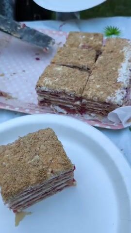 A close-up shot shows a layered cake with frosting and crumbs. A slice of cake is on a white plate in the foreground. The rest of the cake, cut into squares, is on a pink tray in the background. A knife is visible on the tray. The setting appears to be outdoors with a white tablecloth.