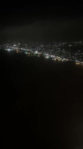 The video is a nighttime aerial view of a coastal city. The city lights are visible along the shoreline, creating a bright band against the dark landscape. The water is dark, reflecting some of the lights. The sky is dark and cloudy. The camera appears to be moving, possibly from an airplane. There are no people or specific objects clearly visible in the frame. The overall scene is serene and captures the beauty of a city at night from above.