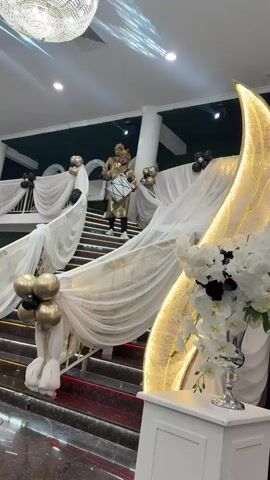 Two men dressed in gold attire, wearing headdresses, stand on a staircase. The man on the left holds a drum in front of him and appears to be playing it. The man on the right also holds a drum and appears to be playing it. Both men are looking forward and slightly upward. The staircase is decorated with white draped fabric and gold and black balloons. A large, illuminated crescent moon decoration is visible to the right of the staircase, with white flowers and black accents in a vase in front of it. The scene appears to be an indoor event, possibly a wedding or celebration, with a large chandelier hanging from the ceiling and speakers visible in the foreground. In the background, a few individuals are seated at tables, and a person is pushing a cart.