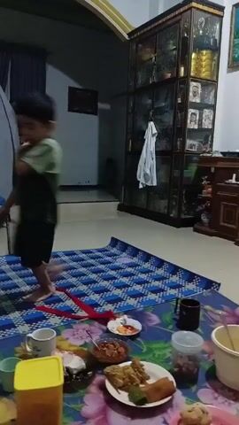 The video shows the interior of a house with a meal laid out on a floral patterned mat on the floor. A young girl in a green outfit runs into the shot, smiling and holding something in her hands. She pauses briefly before another young child, a boy in a similar green outfit, enters the frame and appears to playfully push or chase her. The environment includes tiled floors, wooden furniture, yellow-trimmed pillars, and a staircase in the background.