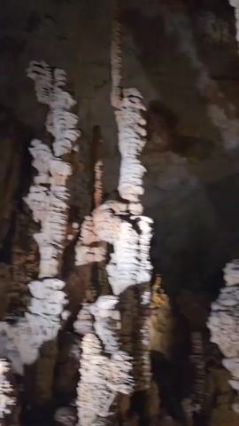 The video shows a dark cave interior with numerous stalactites and stalagmites. The formations are lit by artificial lights, creating a dramatic effect. The camera pans slowly across the scene, highlighting the different shapes and textures of the rock structures. Some of the stalactites are long and slender, while others are thick and columnar. The cave walls are rough and uneven, with visible layers of rock. In some areas, water drips from the ceiling, contributing to the formation of the stalactites and stalagmites. The overall impression is one of natural beauty and geological wonder.