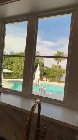 The video shows a view from inside a kitchen, looking out a large window at a backyard with a swimming pool and lush greenery. The kitchen has a marble countertop and a gold faucet. Outside, a bright blue swimming pool is visible, surrounded by a patio. Palm trees and other dense foliage frame the pool area. The sky is a clear blue with scattered white clouds. The camera pans slightly to the right, revealing more of the backyard and then moves to show a hallway leading into a living area. A white oscillating fan stands in the hallway, and the living room appears furnished with a table and chairs.