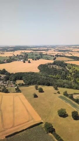 An aerial drone view captures a rural landscape with golden fields and a small village under a clear sky. The fields show tractor tracks, suggesting recent agricultural activity. A cluster of houses with red roofs is visible, nestled amongst trees. The overall scene is peaceful and serene.