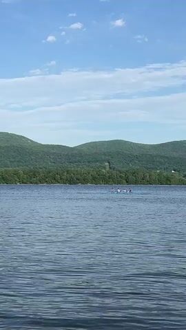 Here's a description of the video:
The camera faces a body of water, rippled by wind. In the background, a mountain range covered in forest is visible under a blue sky with scattered white clouds. A small motorboat, with what appears to be a single occupant dressed in white, moves across the water. Initially, a group of what might be paddleboarders is seen, but they're further away and harder to distinguish.