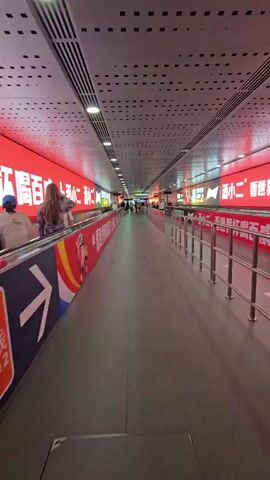 A wide hallway with a moving walkway on the left and a railing on the right is shown. People are walking on the moving walkway and standing. The walls of the hallway are covered in bright red advertisements. The ceiling is made of metal panels with lights integrated into them. The floor is a dark grey tiled surface. The overall impression is one of a modern, busy public space, likely a transportation hub or a large shopping mall.