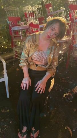 A woman sits on a chair with rows of chairs in the background. She is wearing a gold bomber jacket over a patterned crop top and black wide-leg pants. She is looking down initially, then looks up, smiles, and waves. Her feet are bare except for flip-flops. The background shows rows of transparent chairs stacked on top of each other with red ribbons. The ground appears to be wet, with debris scattered around. Another person's foot, wearing flip-flops, is partially visible in the corner of the frame.
