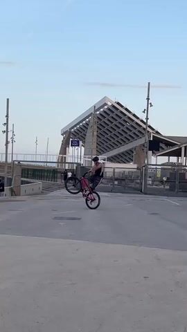 A person wearing a helmet and a patterned shirt is riding a pink bicycle. They approach a small group of people standing with their bicycles. The person on the pink bicycle pops a wheelie, raising the front wheel of the bike off the ground, before riding away. The setting appears to be an outdoor plaza or gathering place with a modern, large structure in the background and a few people walking in the distance. The sky is light blue with a few wispy clouds.