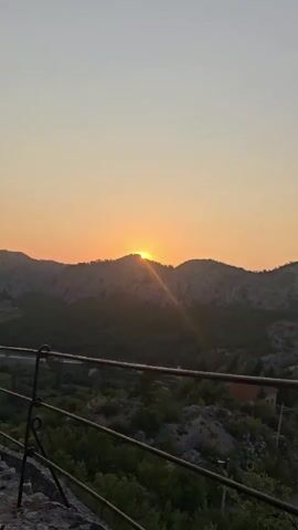 The video shows a time-lapse of a sunset over a mountainous landscape. The sun, a bright orange orb, is partially obscured by the crest of a hill. Rays of sunlight emanate from the sun, casting a warm glow over the scene. The sky is a gradient of orange and yellow, deepening to a soft orange at the top of the frame. The hills are silhouetted against the bright sky, with some sparse vegetation visible on their slopes. The scene is serene and peaceful, capturing the beauty of nature.