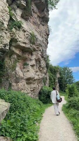 A person in white clothing is walking away from the camera on a dirt path. The path is bordered by lush green vegetation on both sides. To the left, a tall rock formation rises dramatically, covered with patches of greenery. The sky above is a mix of blue and white clouds. The person has a bag slung over their shoulder.