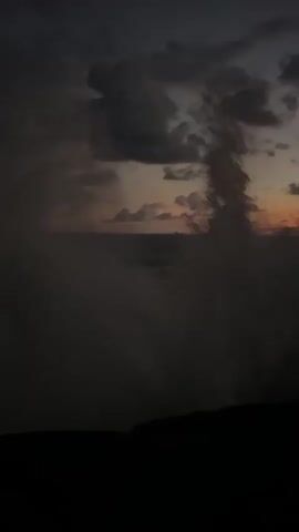 The video captures a dramatic coastal landscape at twilight, likely either sunrise or sunset, based on the vibrant colors on the horizon. The scene unfolds from a close, dark view, then pans up and out to reveal a vast expanse of ocean. In the immediate foreground, dark, rugged rock formations are visible, with gentle waves crashing against them.

The sky is a prominent feature, dominated by heavy, dark grey clouds that span across the upper part of the frame, creating a moody atmosphere. Below these clouds, the horizon glows with intense shades of orange, red, and yellow, painting a striking contrast with the darker sky and sea.

No people or man-made objects are visible, emphasizing the natural beauty and solitude of the location. The overall ambiance suggests a powerful yet serene natural setting. The location is consistent with a coastal area such as Rumassala, Sri Lanka. There is no visible text in the video.