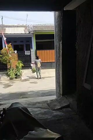 A person wearing camouflage pants, a light-colored backpack, and a dark shirt stands in the middle of a street, holding a white object. They are facing away from the camera and towards two houses. The house on the left is painted blue and has a white-striped roof. A flagpole with an Indonesian flag is planted in front of it. The house on the right has an orange-brown facade and a black gate. Green plants are visible in pots near the blue house. The street is paved. A dark-colored object, possibly a motorcycle cover, is in the foreground. The scene appears to be outdoors on a sunny day.