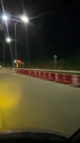 A dashcam perspective captures a nighttime scene from a vehicle driving on a paved road. The road has two lanes, separated by a solid white line, and is illuminated by streetlights on tall poles. On the right side of the road, there's a barrier made of red and white construction barriers. Two traffic signs are visible: a blue circular sign with a white arrow pointing right and down, indicating a mandatory turn, and a red circular sign with a white horizontal bar, indicating "do not enter" or "wrong way." In the distance, an oncoming white semi-trailer truck approaches, its headlights shining brightly. As the video progresses, the truck passes by on the left side, moving in the opposite direction. The sky is dark, typical of nighttime.