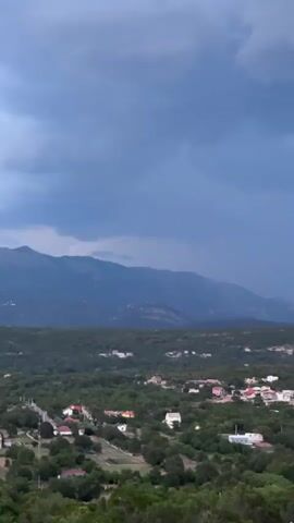 This is a landscape video of a rural village nestled in a valley with mountains in the background. The village consists of numerous houses with red and white roofs, scattered amongst lush green trees. A road winds through the village. The scene is set under a dramatic, overcast sky with dark, heavy clouds suggesting an impending storm. The mountains in the distance are shrouded in a blue haze, their peaks obscured by the low-lying clouds. The overall atmosphere is moody and somewhat somber due to the weather. The lighting indicates it is either early morning or late evening, as the light is dim and diffused. There are no people or specific objects clearly identifiable in the foreground or midground. The video seems to capture a moment of transition, with the natural beauty of the landscape juxtaposed against the dramatic weather.