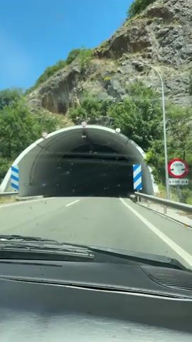 The video begins from the perspective of someone inside a vehicle, looking forward through the windshield at the entrance of a tunnel. The tunnel has a white, arched ceiling and concrete walls. There are signs visible at the entrance indicating height restrictions. As the vehicle enters the tunnel, the lighting changes from bright daylight to artificial lights lining the tunnel walls. The tunnel walls are a mix of rough-hewn rock and smoother, panelled sections, some of which are illuminated with blue lights. The road surface is asphalt with white lane markings. The vehicle's windshield wipers are visible in the foreground. The camera continues to move through the tunnel, showing other vehicles in the distance. The tunnel curves gently to the right as it progresses. The video ends with the view still inside the tunnel, with artificial lights illuminating the path ahead.