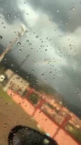 The video shows a rain-covered window with a partial rainbow visible through it. The sky is overcast and grey. A blurred orange structure is visible through the window in the lower portion of the frame. The camera angle is tilted slightly upward.