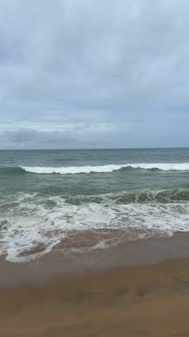 The video captures a close-up view of a beach on a cloudy day. The foreground consists of wet sand, showing the receding tide. In the middle ground, gentle waves crash onto the shore, creating white foam. The ocean extends into the background, with the horizon line dividing it from a cloudy sky. The overall color palette is muted, dominated by grays, blues, and browns. There are no people or specific objects visible in the frame.