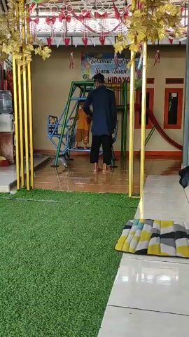 A person in dark pants and a dark jacket stands with their back to the camera, watching a child on a swing set. The child is wearing an orange shirt and dark pants. The swing set is green and blue. A banner with the words "Selamat Datang Siswa Siswi" and "HIDAYAH" is visible behind the swing set. The environment appears to be an outdoor area with artificial grass in the foreground and a tiled floor in the background. There are also decorative gold columns with leaf accents.