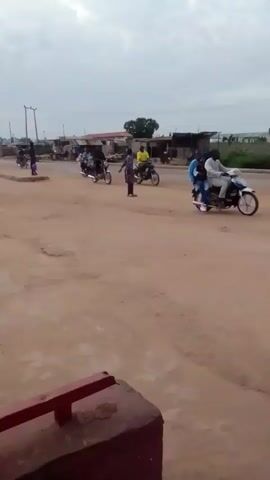 The video shows a dusty, unpaved road with several motorbikes and a three-wheeled auto-rickshaw. In the foreground, a red metal railing is visible. In the background, there are some low-rise buildings and utility poles. Two people are riding on a motorbike in the middle ground. Another person, dressed in a yellow shirt, is riding a motorbike to the right. A person dressed in a purple and white garment is standing near the center of the road. A green auto-rickshaw with two people inside drives from left to right across the frame. The sky is overcast.