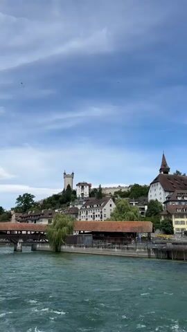 A wide shot shows a river flowing in the foreground with a town built on a hill in the background. The town has traditional European architecture, with terracotta roofs and white walls. A prominent castle-like structure stands on the highest point of the hill. A covered bridge spans a section of the river in the midground. The sky is blue with scattered white clouds. The water in the river is a vibrant turquoise color and appears to be moving with some current. The camera pans slightly to the right, revealing more of the town and its buildings. There are trees scattered throughout the town, and a very tall, slender tree is visible on the far right of the frame. The scene is tranquil and picturesque.