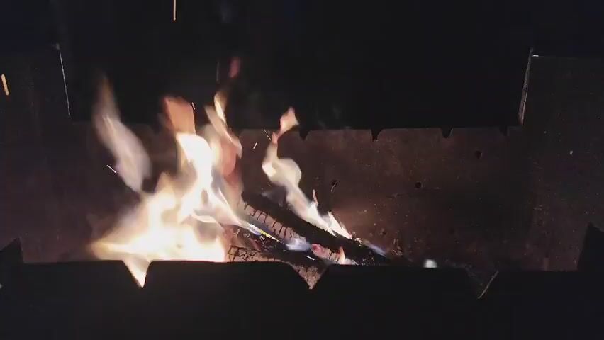 A close-up shot shows burning wood inside a metal grill. The flames are orange and yellow, with hints of blue at the base. Smoke rises from the fire, and embers occasionally spark and fly upwards. The background is dark, suggesting nighttime or a poorly lit environment. The grill appears to be made of dark metal and has a serrated edge along the top. The logs within the grill are partially consumed by the fire.