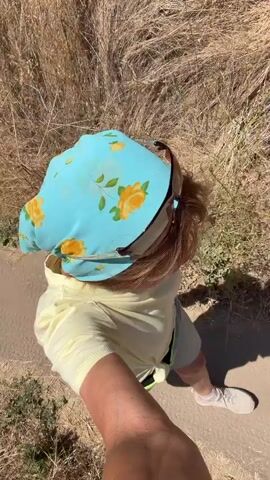 A video captures a person from a first-person perspective, looking down at the person's back. The person is wearing a light yellow hoodie, light-colored shorts, white sneakers, and a light blue bandana with yellow flowers. The person is holding a dark object, possibly a phone, in their right hand, and is walking on a dirt path with dry grass on either side. The sun casts a shadow of the person and an object in their hand onto the path. The scene appears to be outdoors, possibly in a dry, grassy area.