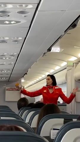 A flight attendant in a red uniform with a red scarf is demonstrating the use of an oxygen mask on an airplane. She is holding an orange oxygen mask and is standing in the aisle between rows of seats, facing towards the front of the plane. Passengers are visible in the foreground, seated and looking towards the flight attendant. The overhead compartments and an illuminated exit sign are visible above.
