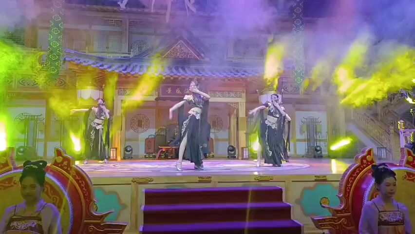 A group of women in elaborate black and gold costumes are performing a dance on a stage decorated with traditional Asian architecture. The stage is lit with colorful spotlights and smoke effects. The dancers have their hair styled in elaborate updos. Two women in similar costumes are also visible on either side of the stage, appearing to be part of the set or audience. The overall atmosphere suggests a theatrical performance.