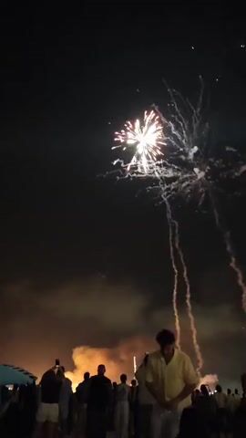 This media file captures a lively fireworks display at night, likely on a warm evening given the casual attire of the crowd. The scene is set outdoors, with a dark night sky serving as the canvas for the vibrant pyrotechnics. Silhouettes of people are visible in the foreground, many holding up their phones to capture the spectacle, indicating a shared experience and excitement.

The fireworks are the central focus, exploding in bursts of white, pink, and red light, creating dramatic streaks and patterns against the darkness. Trails of smoke descend from the explosions, adding to the atmospheric quality of the display. The presence of a beach umbrella on the far left suggests a coastal location, and the "Valdelagranas, Spain" context further pinpoints it to the beach area of Valdelagrana.

The overall mood is celebratory and festive, typical of events like national holidays or special occasions. The bright lights of the fireworks contrast with the dark sky and the silhouettes of the onlookers, emphasizing the ephemeral beauty of the display. No visible text is present in the media file.