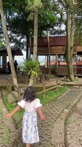 A young girl in a light pink shirt and patterned blue and pink dress is walking on a pathway through an outdoor area. The path is made of grey blocks spaced apart, with small grey stones lining either side. A short wooden fence runs along the left side of the path, and there are several trees scattered throughout the scene, some of which line the path. Several small wooden buildings with reddish-brown roofs are visible through the trees. Other people are visible in the background around the buildings. The girl raises her hands up in the air. The weather appears overcast.
