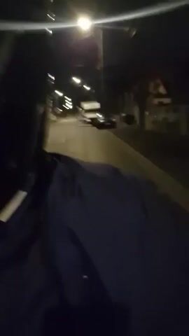 A first-person perspective video shows a person riding a motorcycle down a dark residential street at night. The person is wearing a black helmet and a blue jacket. The street is lit by streetlights. There are houses and parked cars visible on either side of the street. The camera is shaky, likely due to the movement of the motorcycle.