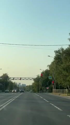A vertical, low-angle shot captures a wide asphalt road lined with lush green trees on either side. The sky above is a clear, pale blue, transitioning to a lighter shade near the horizon. Overhead, power lines stretch across the frame. Several cars are visible on the road, moving away from the camera, while others are stationary at a traffic light. Traffic lights, illuminated in red, are prominently displayed, indicating that traffic is stopped. A pedestrian crossing sign is also visible. The scene appears to be a sunny day on a suburban or rural road.