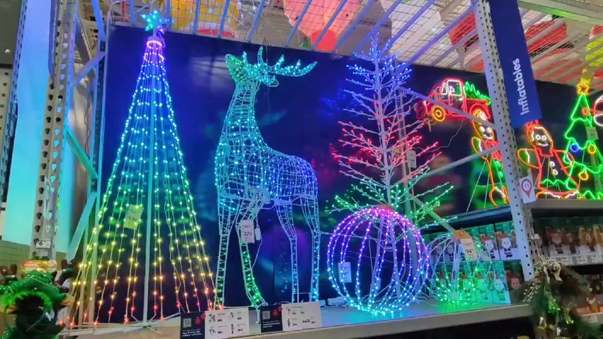The video shows a display of illuminated Christmas decorations inside what appears to be a store. The decorations include a cone-shaped Christmas tree, a reindeer, a bare tree, and a sphere, all made of wireframes covered with colorful LED lights. Each item has a price tag attached. Behind the decorations is a backdrop depicting additional Christmas-themed figures, also illuminated. Above the display is a complex network of metal supports, typical of store shelving. A sign labeled "Inflatables" is visible on the right side of the frame. The camera angle is static, providing a clear view of the entire arrangement of decorations.