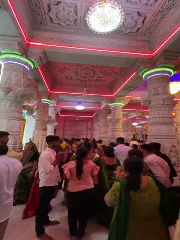 The video shows a crowded temple interior with white marble columns, a carved ceiling, and colorful LED lighting. People of various ages and ethnicities are seen walking and standing, some with their hands clasped in prayer. The columns are decorated with green and blue lights, and the ceiling has red neon strips. A chandelier hangs from the center of the ceiling. The atmosphere is lively and appears to be during a religious event.