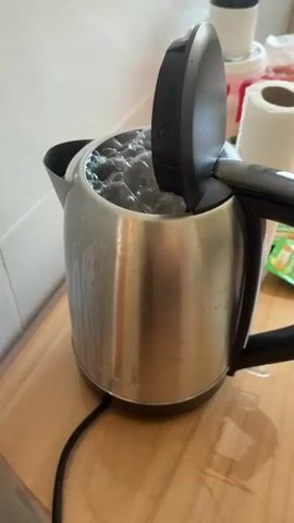 The video shows a person preparing pasta in a kettle. The kettle is stainless steel and sits on a light wooden countertop. The lid is open, revealing boiling water with what appears to be brown pasta inside. The countertop is slightly wet, with a black power cord visible next to the kettle. Other items, including a white container with a red label, a roll of paper towels, and a box of tea bags, are visible in the background. The tiles of the backsplash are white.