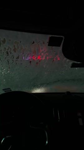 The view from inside a car at a car wash. Bubbles and soapy water cover the windshield. Outside the windshield, colored lights flash. The steering wheel is visible in the dark car interior. No people are visible.