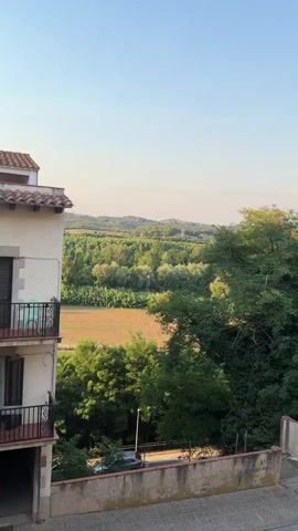 The video begins with a view of a building with a tiled roof and a balcony overlooking a rural landscape. The landscape consists of rolling hills covered in lush green trees and fields of golden crops. In the distance, there are some buildings and power lines. The camera pans across the scenery, revealing more of the natural beauty. As the video progresses, the view shifts to a road with a sidewalk and streetlights. The road is lined with trees and a low concrete wall. The sky is a clear blue, with a few wispy clouds. The overall impression is one of a peaceful and idyllic countryside setting.