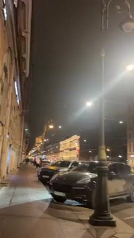 The video shows a street scene at night, illuminated by streetlights. The street is lined with parked cars, including a Porsche Cayenne, and several people are walking on the sidewalk. The buildings on either side of the street are tall and well-lit.