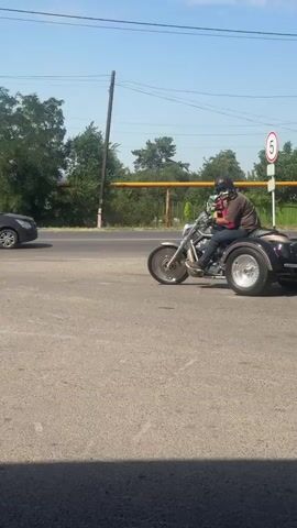 A person wearing a helmet and goggles rides a black and silver trike on a paved road. The trike has two wheels in the back and one in the front. The rider is wearing a dark t-shirt, blue jeans, and gloves. The trike is moving from left to right in the frame. In the background, there are trees, a telephone pole, a gray sedan, a blue car, a white van, and a building with a dark roof. The sky is blue and clear.
