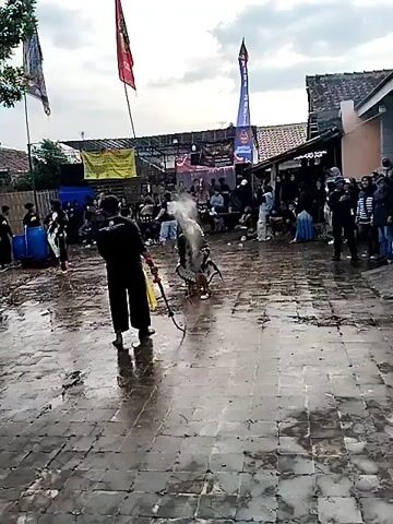 The video shows an outdoor event with several people and a wet, paved ground. In the foreground, a person in black is holding a whip with a red tip. Two performers in elaborate costumes are engaged in a dance or fight. One performer wears a black and white outfit with red fringes and a mask, while the other wears a dark costume and has a whip. Other people are watching the performance from behind a barrier. There are banners and flags in the background, suggesting a festive or cultural gathering. The overall atmosphere appears dynamic, with water splashing and performers moving energetically.