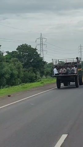 A trailer pulled by a tractor is moving down a paved road. Several people are sitting in the open back of the trailer. The trailer is green with yellow accents. A tall metal tower with wires is visible in the background, along with trees and a cloudy sky.