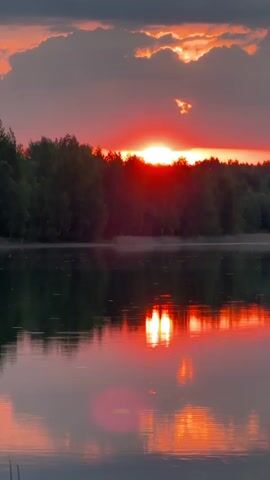 The video shows a sunset over a lake. The sun is setting behind a treeline, and the sky is filled with red and orange clouds. The lake is calm and reflects the colors of the sky. There is a blurry red circular shape reflected in the lake.