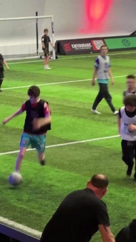 The video shows a group of people watching an indoor soccer game. The game is being played on a green artificial turf field with white lines. Several players, some wearing vests, are visible on the field. Some spectators are standing close to the field, observing the game and occasionally talking. At one point, some are seen holding drinks. The setting appears to be a sports complex or entertainment venue. Logos and advertisements can be seen on the surrounding walls.