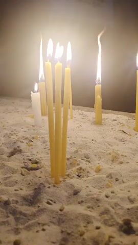 Several lit candles stand in a bed of sand. The candles are of varying heights and thicknesses, with some taller, thinner yellow candles grouped together in the foreground. Other candles, including a white one and several yellow ones, are scattered throughout the scene. The flames of the candles flicker, casting a warm glow on the sand and the dark background. A plaque with text and a cross is visible on the wall in the upper portion of the frame. The overall impression is of a solemn or devotional setting.
